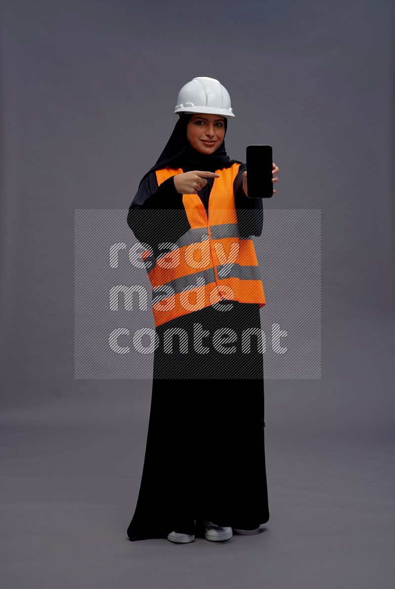 Saudi woman wearing Abaya with engineer vest standing showing phone to camera on gray background