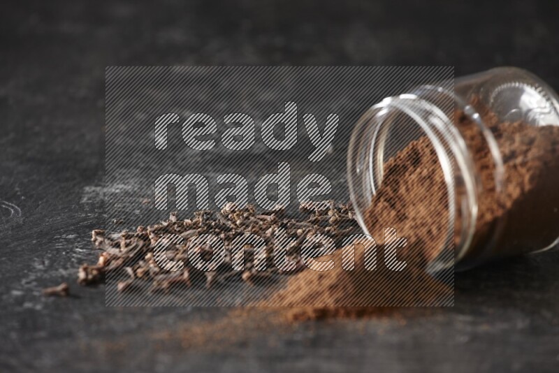 A flipped glass jar full of cloves powder with cloves spread on a textured black flooring