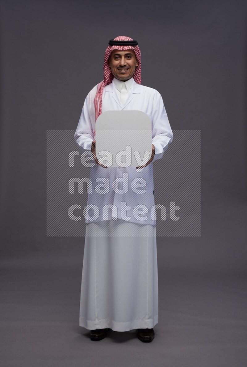 Saudi man wearing thob with lab coat and shomag with pocket employee badge standing holding social media sign on gray background