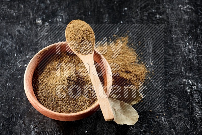 A wooden bowl and spoon full of cumin powder on a textured black flooring