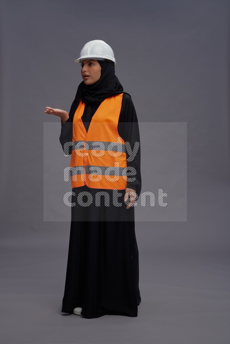 Saudi woman wearing Abaya with engineer vest standing interacting with the camera on gray background