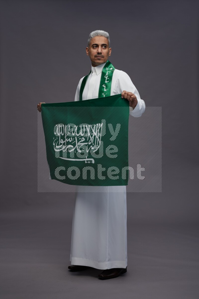 Saudi man wearing thob standing holding Saudi flag on gray background