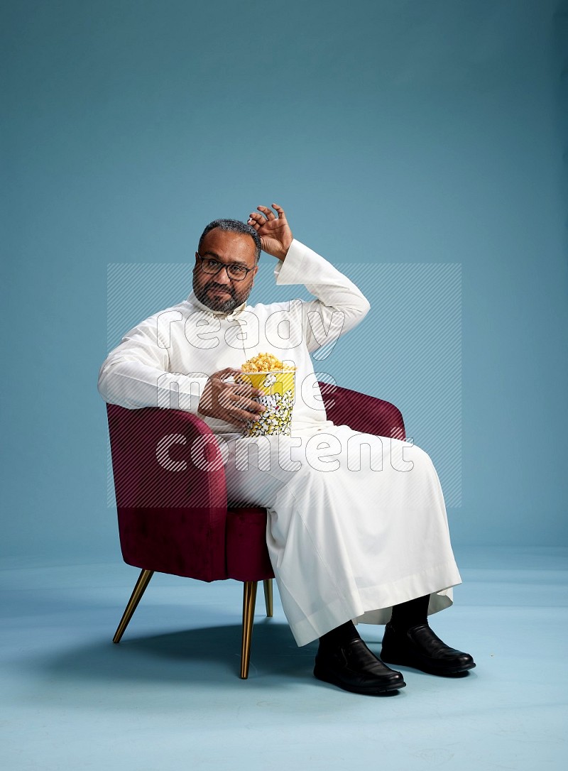 Saudi Man without shimag sitting on chair eating popcorn on blue background