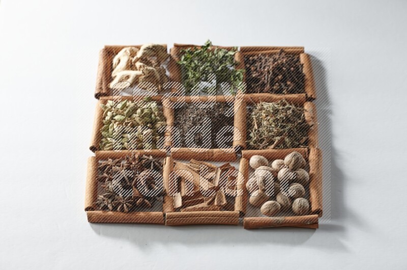 9 مربعات من أعواد القرفة ممتلئة بالشاي في المنتصف محاطة بجوزة الطيب والنعناع المجفف والقرنفل والريحان المجفف والزنجبيل المجفف والقرفة واليانسون والهيل على أرضية بيضاء