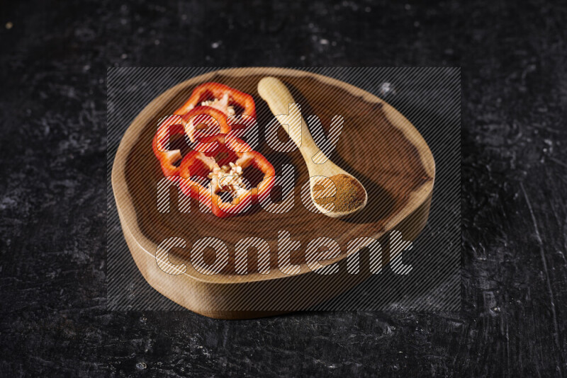 A wooden spoon full of ground paprika powder with red bell pepper slices beside it, all on a wooden tray on black background