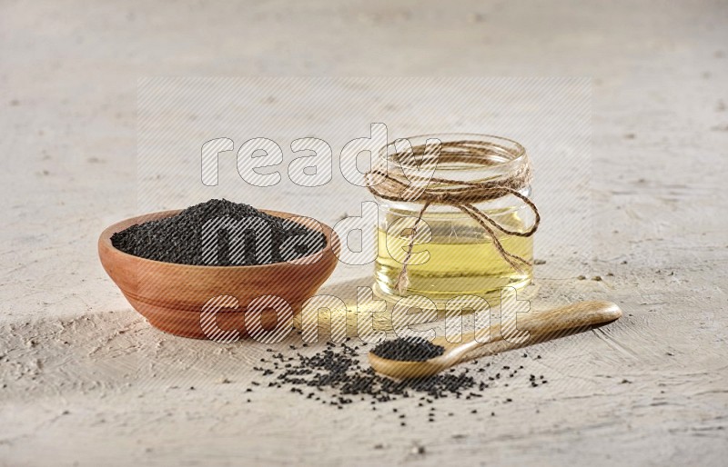 A wooden bowl and spoon full of black seeds with a glass jar of black seeds oil on a textured white flooring