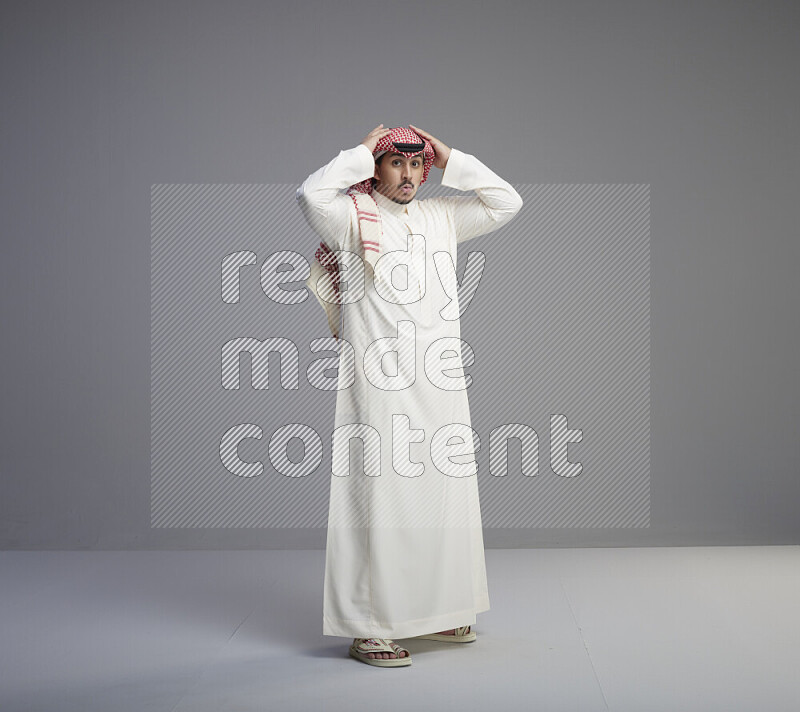 A Saudi man standing wearing thob and red shomag interacting with the camera on gray background