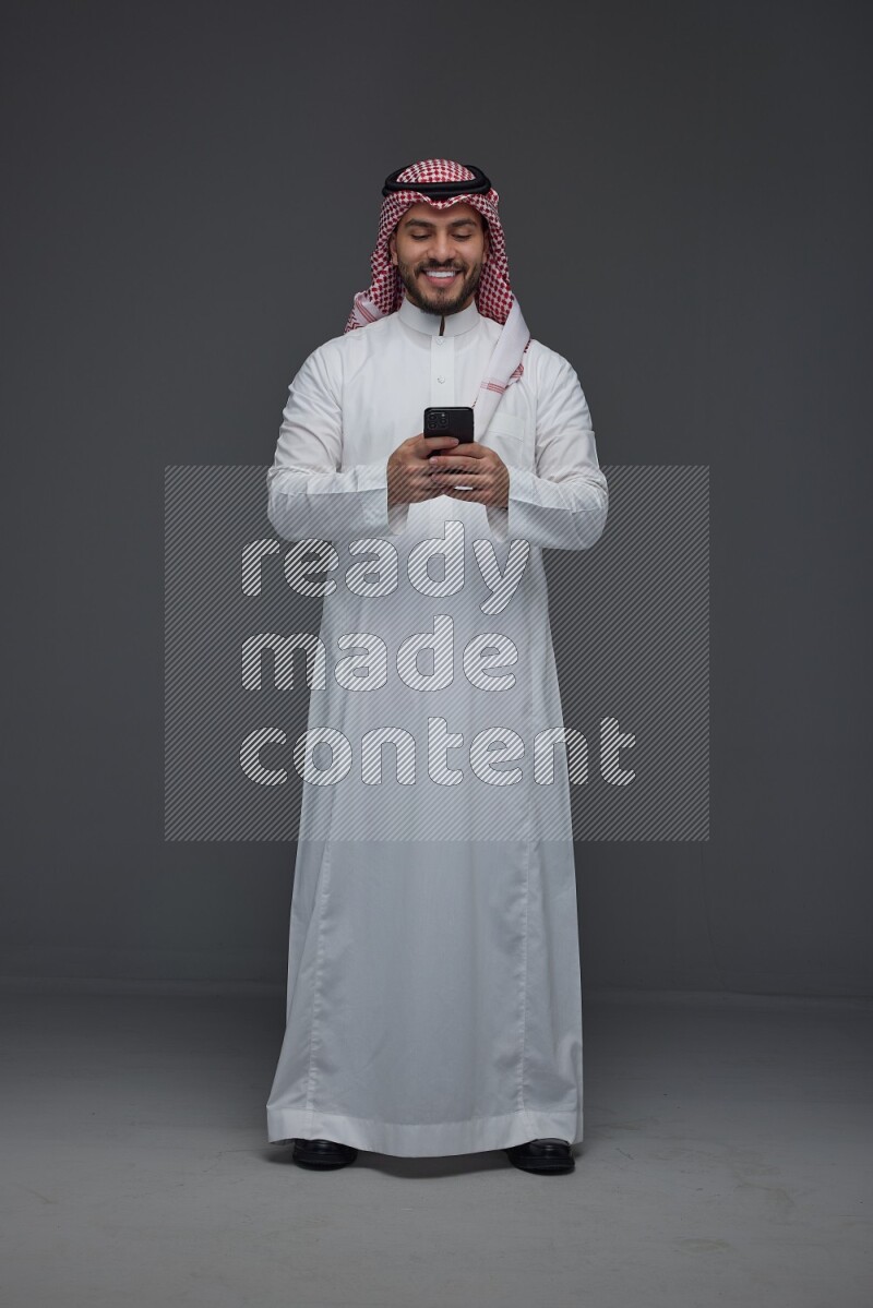 A Saudi man wearing Thobe and Shmagh standing and using his phone eye level on a gray background