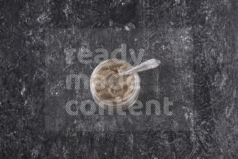 بودرة الفلفل الأسود في وعاء زجاجي مع ملعقة معدنية علي خلفية سوداء