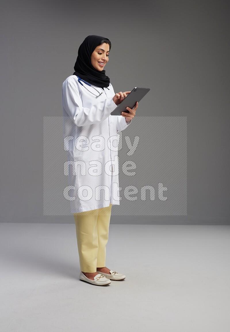 Saudi woman wearing lab coat with stethoscope standing working on tablet on Gray background