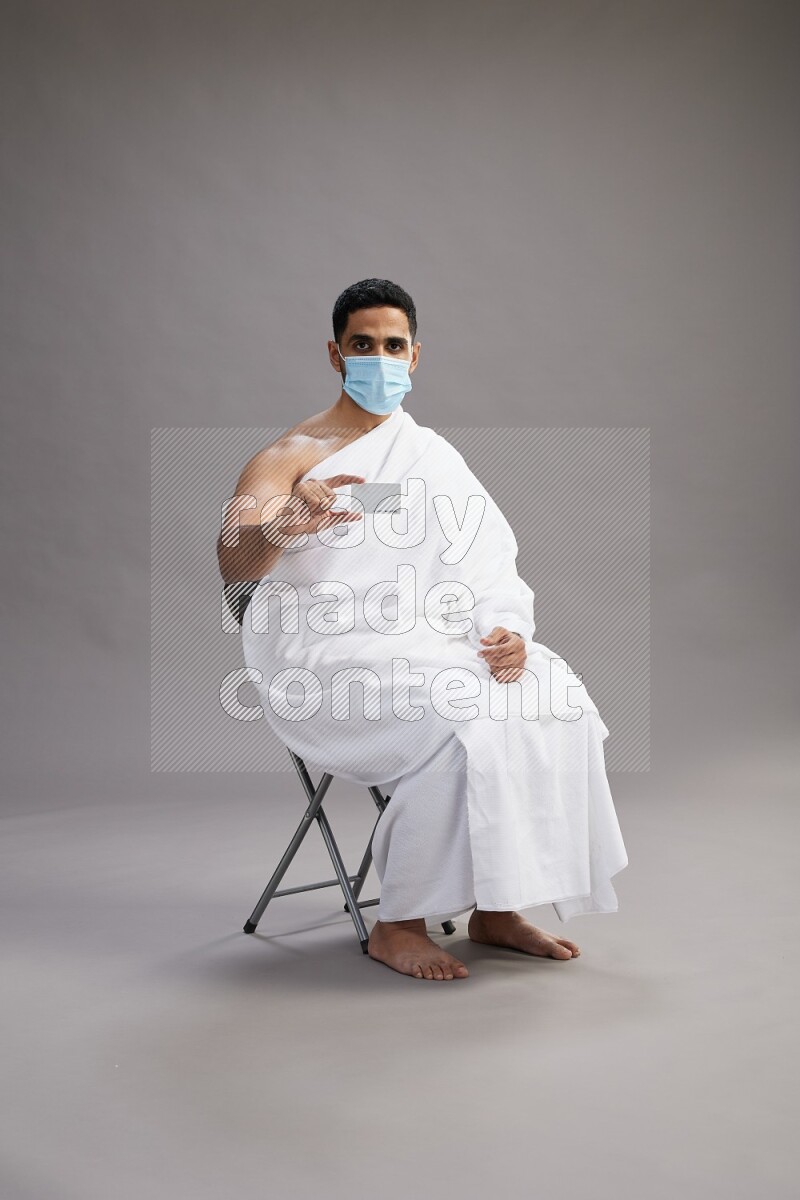 A man wearing Ehram with face mask sitting on chair holding ATM card on gray background