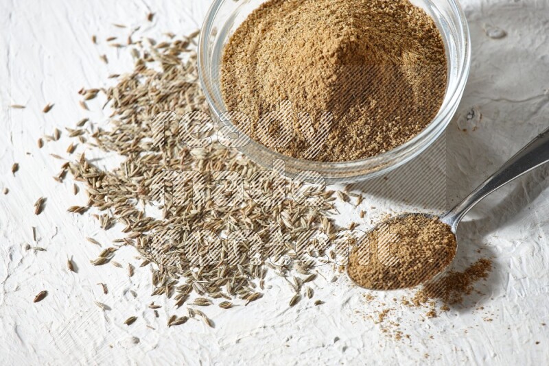 A glass bowl and metal spoon full of cumin powder and cumin seeds underneath it on textured white flooring