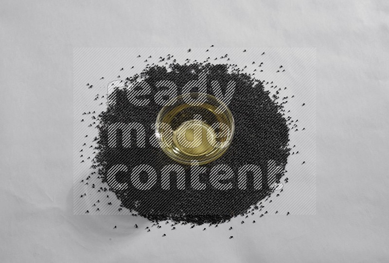 A glass bowl full of black seeds oil surrounded by the seeds on a white flooring