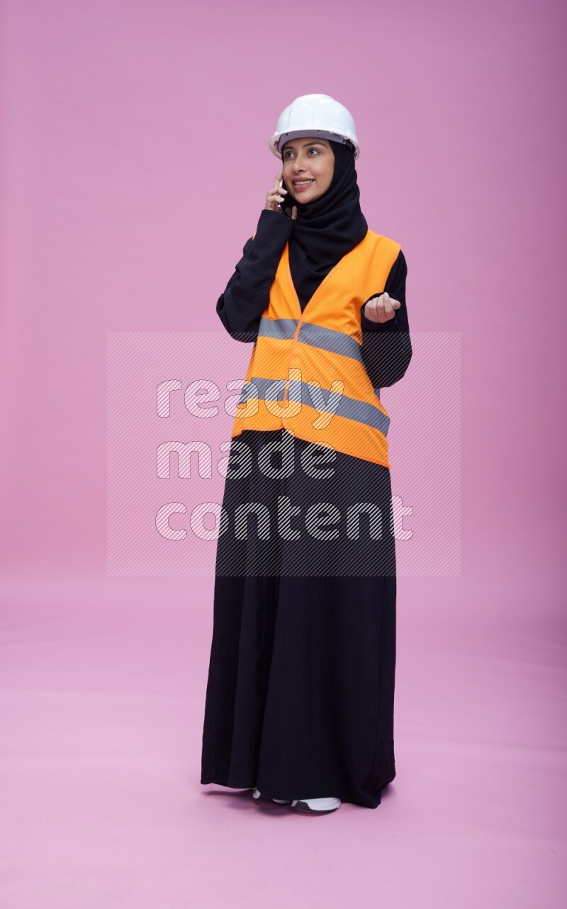 Saudi woman wearing Abaya with engineer vest and helmet standing talking on phone on pink background