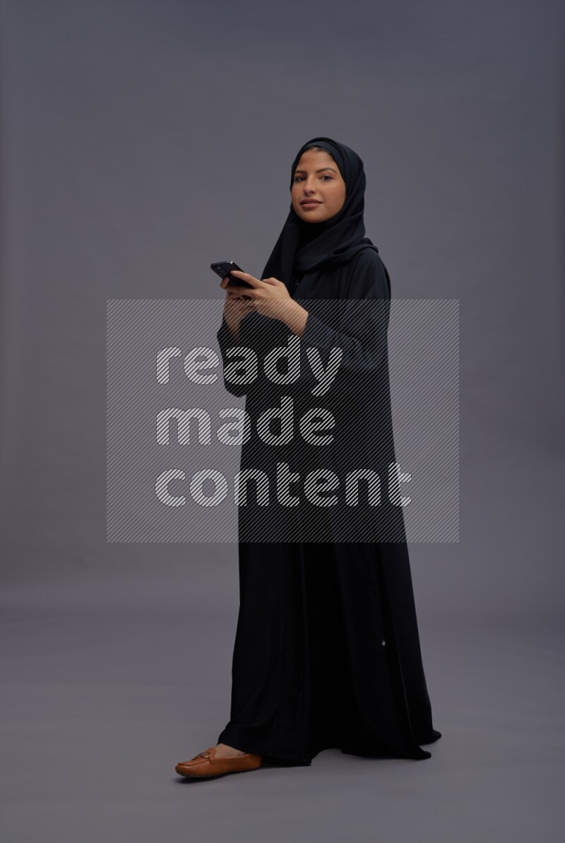 Saudi woman wearing Abaya standing texting on phone on gray background
