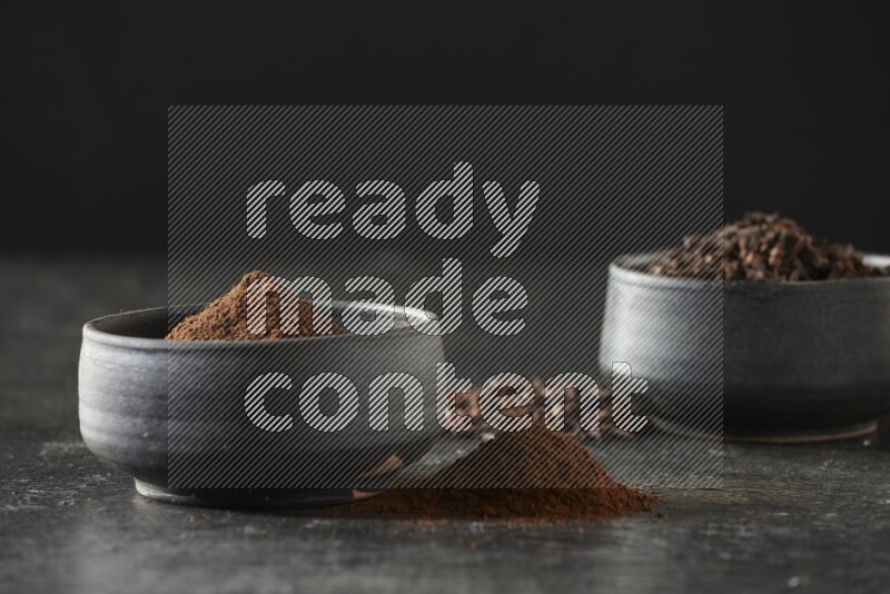 2 Black pottery bowls full of cloves and the other full of cloves powder on textured black flooring