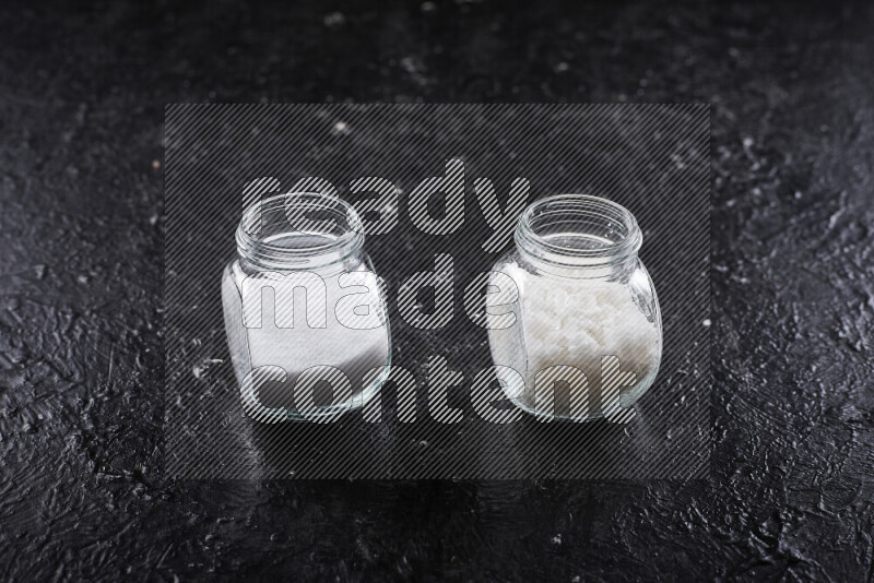 2 glass jars one is filled with table salt and the other with coarse sea salt on black background