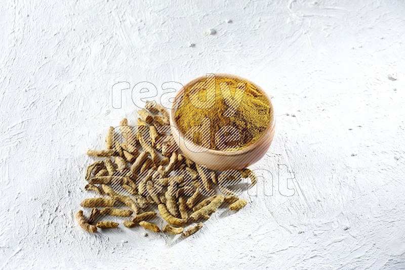 A wooden bowl full of turmeric powder and dried whole fingers beside it on a textured white flooring