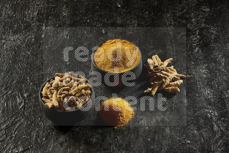 2 pottery black bowls, one full of turmeric powder and the other full of dried turmeric whole fingers on a textured black flooring