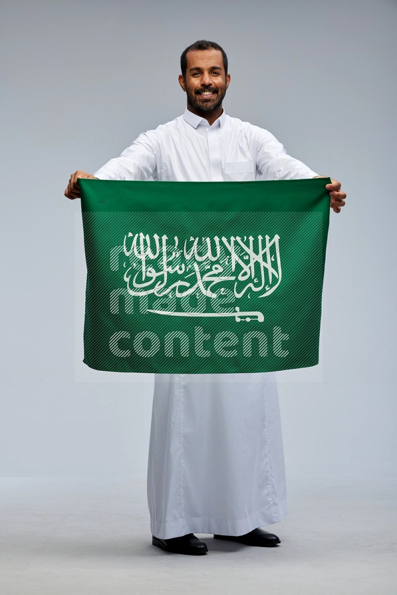 Saudi man wearing Thob standing holding Saudi flag on Gray background