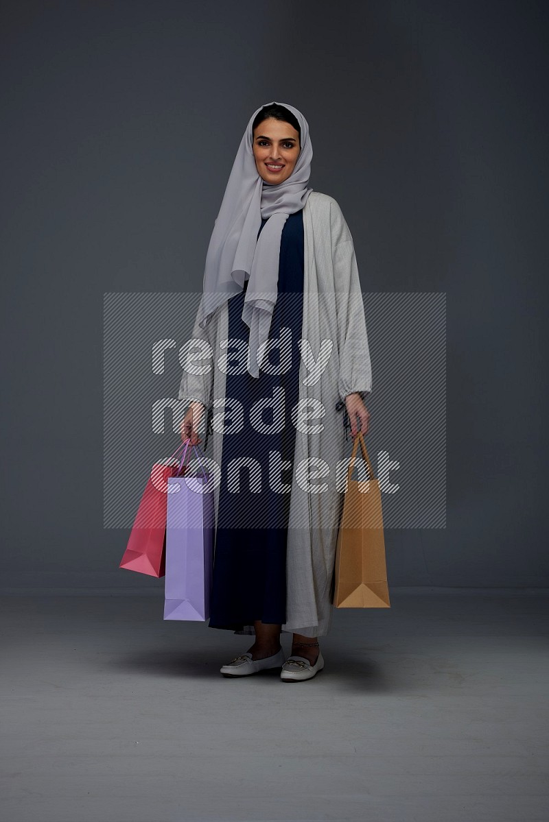 A Saudi woman wearing a light gray Abaya and head scarf standing and holding shopping bags on a grey background