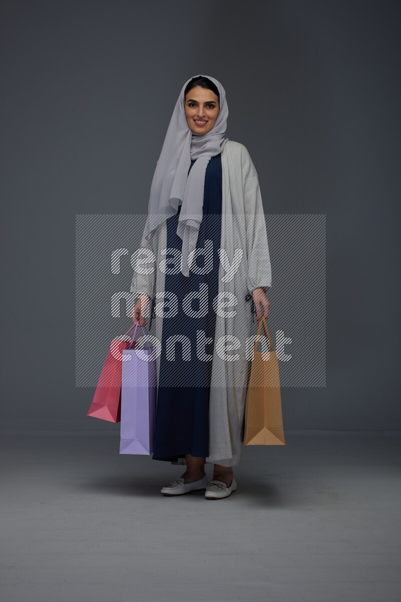 A Saudi woman wearing a light gray Abaya and head scarf standing and holding shopping bags on a grey background