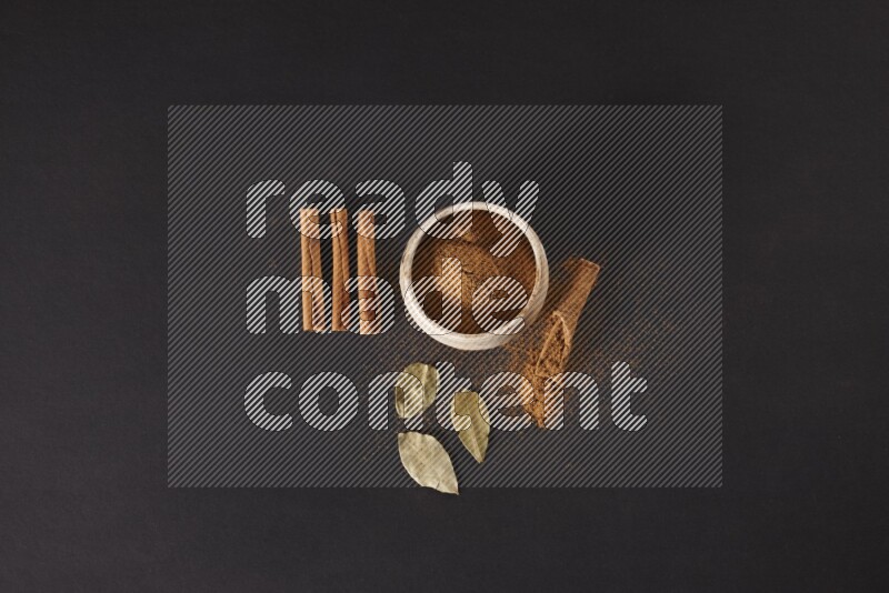 Cinnamon powder in a white pottery bowl and cinnamon sticks and laurel leaves on black background