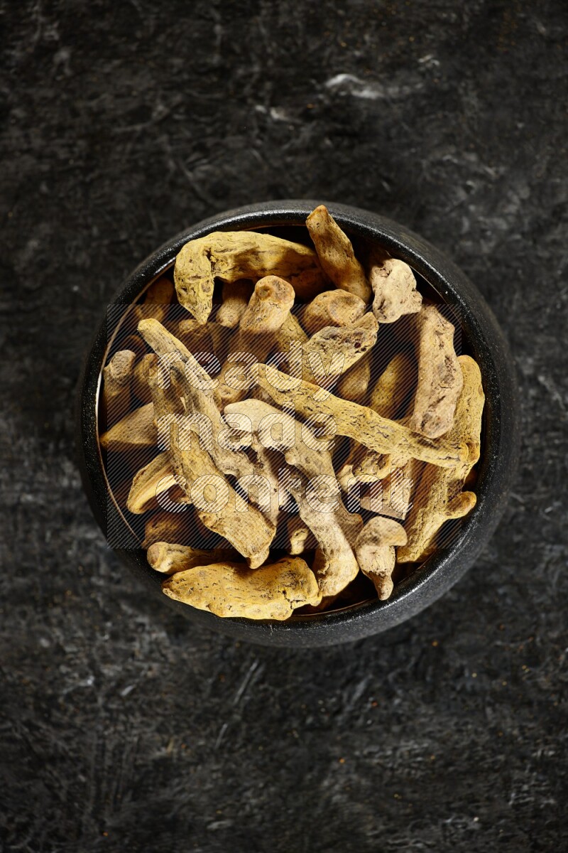 A black pottery bowl full of dried turmeric whole fingers on textured black flooring