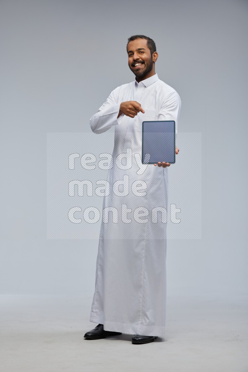 Saudi man wearing Thob standing showing tablet to camera on Gray background
