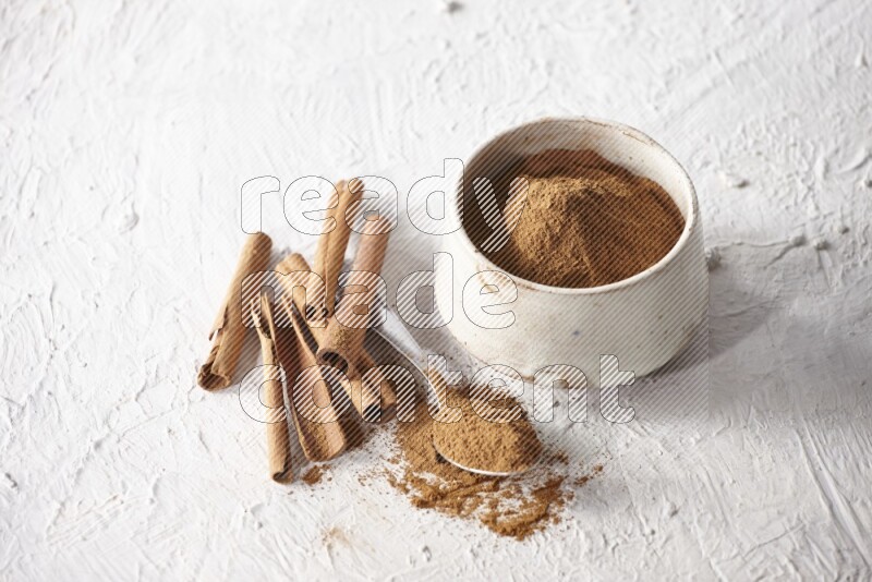 Ceramic beige bowl full of cinnamon powder and a metal spoon with cinnamon sticks next of it on a textured white background