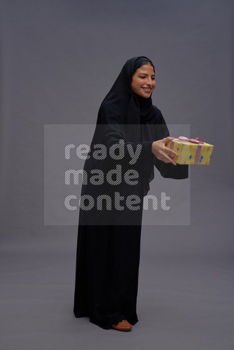 Saudi woman wearing Abaya standing holding gift box on gray background