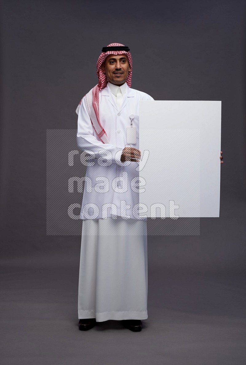 Saudi man wearing thob with lab coat and shomag with pocket employee badge standing holding board on gray background