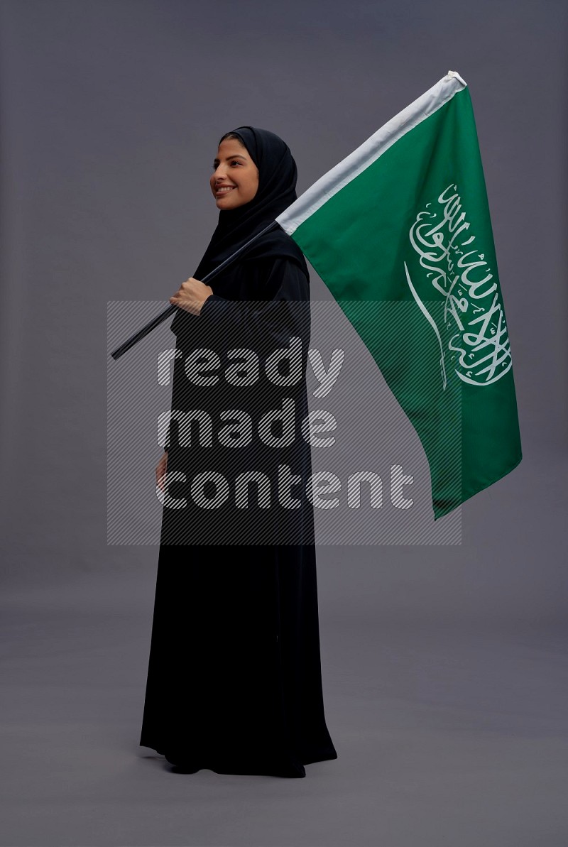 Saudi woman wearing Abaya standing holding Saudi flag on gray background