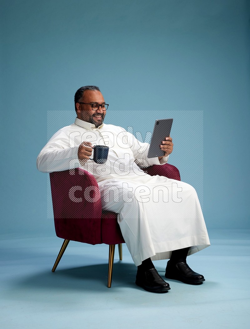 Saudi Man without shimag sitting on chair working on tablet on blue background