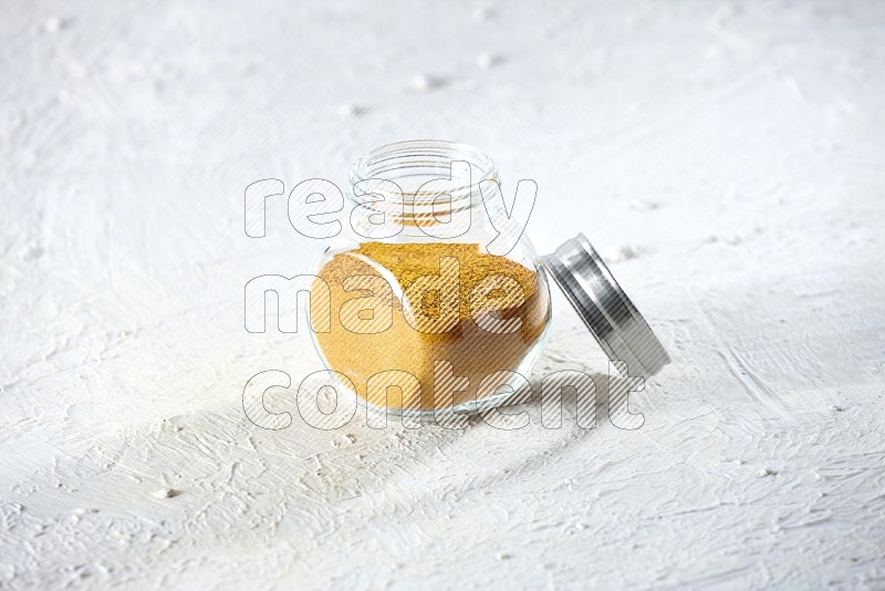 A glass spice jar full of turmeric powder on a textured white flooring