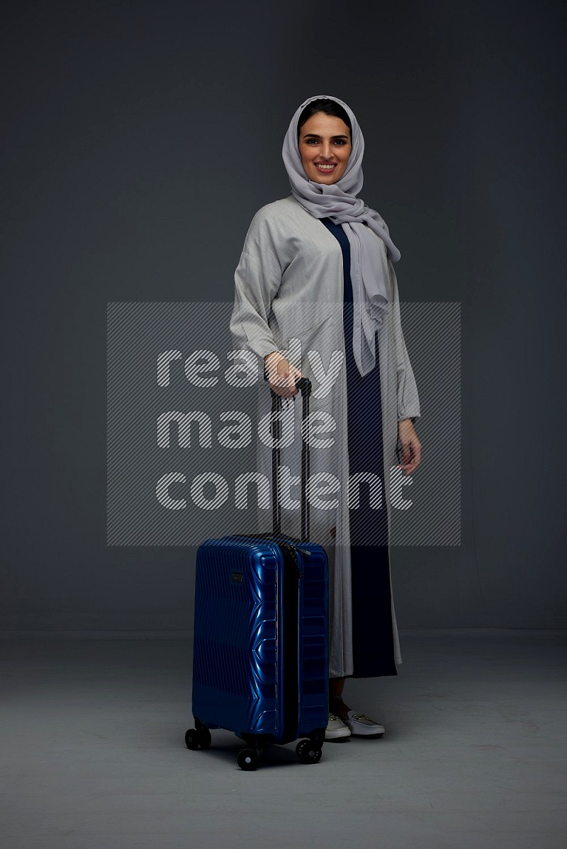 A Saudi woman wearing a light gray Abaya and head scarf standing and showing the tablet's screen eye level on a grey background