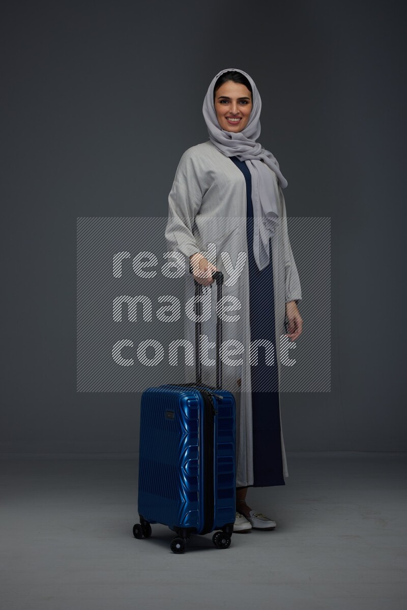 A Saudi woman wearing a light gray Abaya and head scarf standing and showing the tablet's screen eye level on a grey background