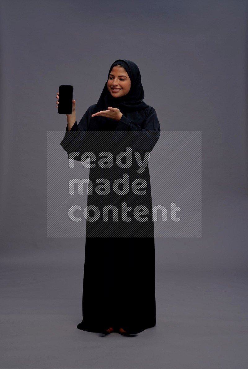 Saudi woman wearing Abaya standing showing phone to camera on gray background