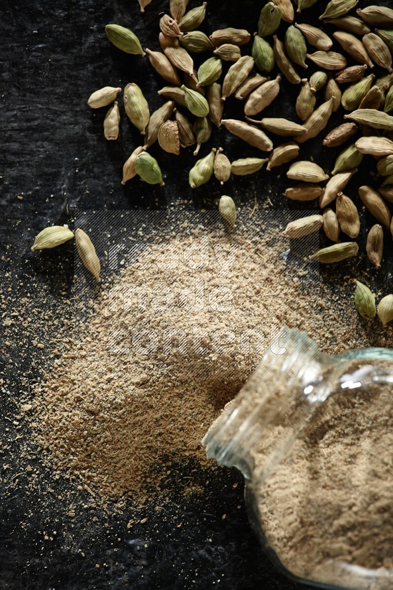 A flipped glass spice jar full of cardamom powder and powder spilled out of it with cardamom seeds spreaded on textured black flooring