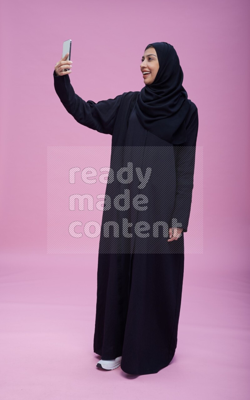 Saudi woman wearing Abaya standing taking selfie on pink background