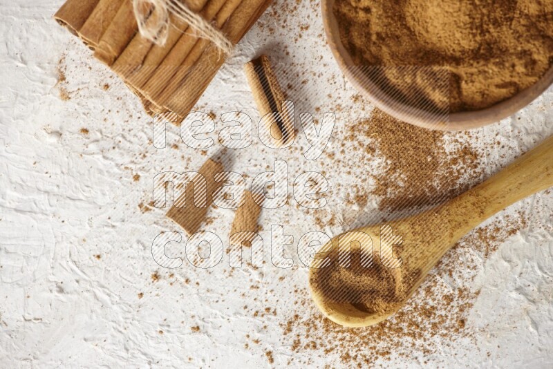 Cinnamon sticks stacked and bounded beside a wooden bowl full of cinnamon powder and a wooden spoon full of powder on white background
