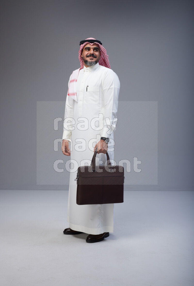 Saudi man Wearing Thob and red Shomag standing holding bag on Gray background