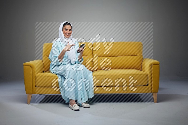 A Saudi woman wearing a light blue Abaya and a white head scarf sitting on a yellow sofa and holding her phone while pointing to it eye level on a grey background