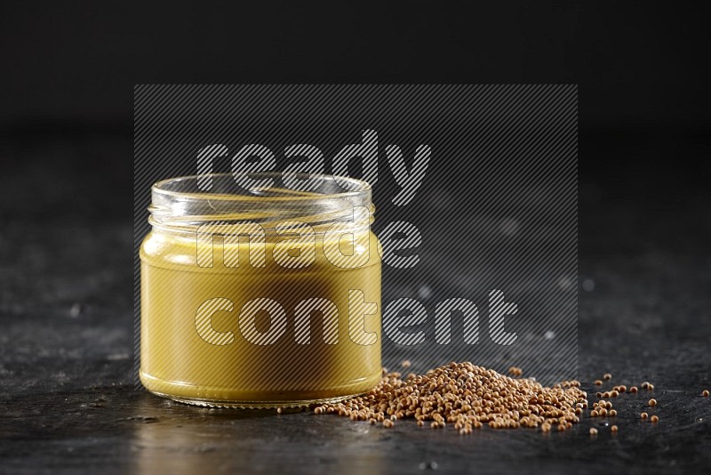A glass jar full of mustard paste and mustard seeds spread next to it on a textured black flooring in different angles