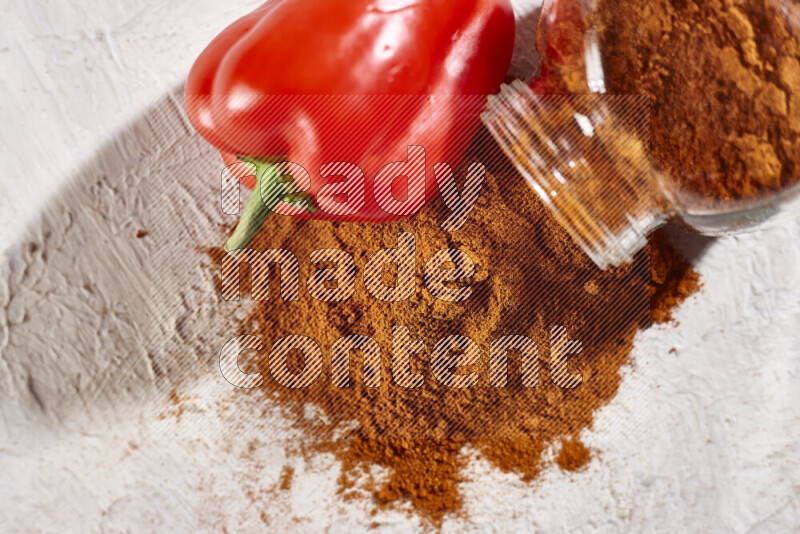 A glass jar full of ground paprika powder flipped with some spilling powder on white background