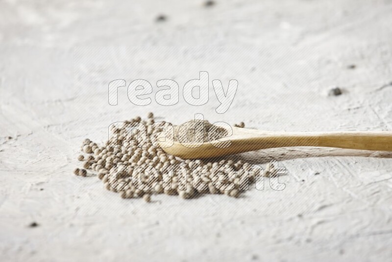 A wooden spoon full of white pepper powder and white pepper beads on textured white flooring