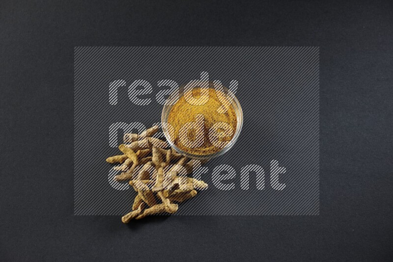 A glass bowl full of turmeric powder and dried whole fingers beside it on black flooring