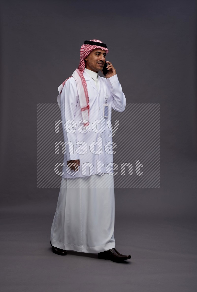 Saudi man wearing thob with lab coat and shomag with pocket employee badge standing talking on phone on gray background