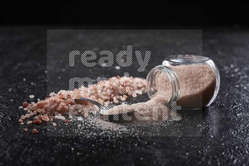 A glass jar full of fine himalayan salt with some himalayan crystals beside it on a black background