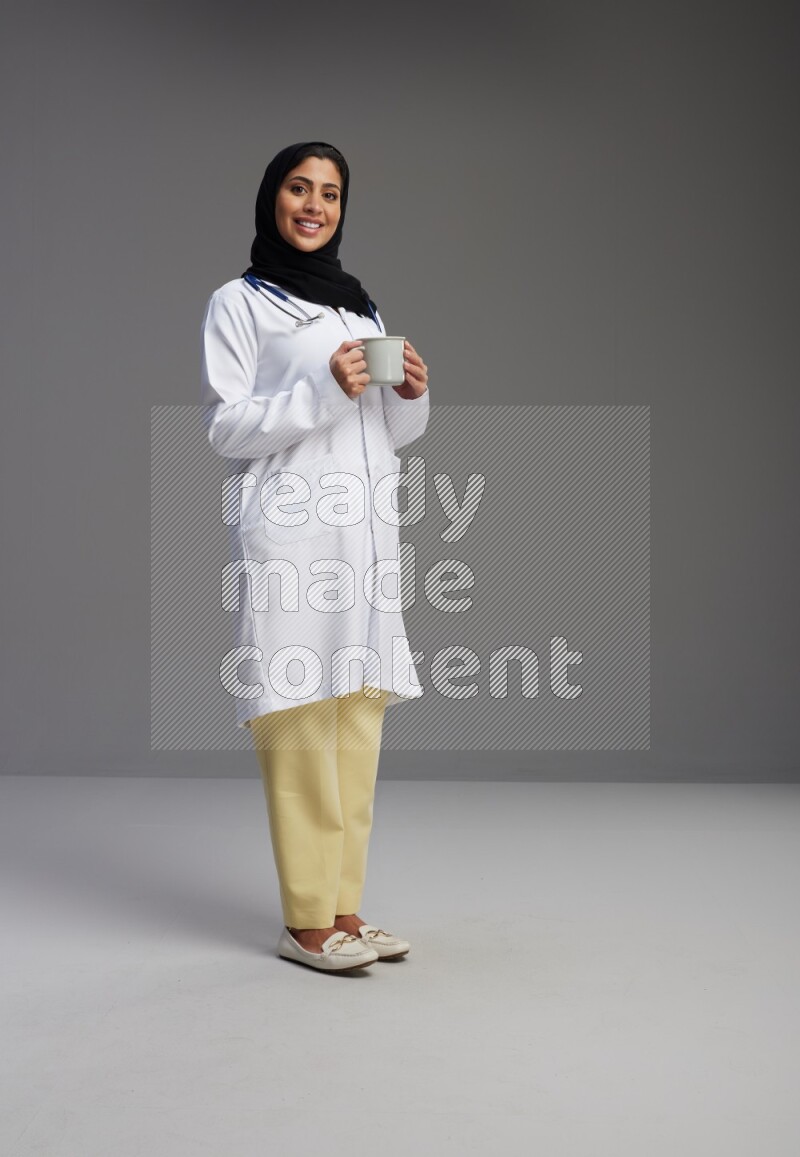 Saudi woman wearing lab coat with stethoscope standing holding mug on Gray background